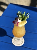 A refreshing tropical cocktail is served in a stemmed glass on a blue table. The drink features a frothy orange concoction and is garnished with a sprig of fresh mint, a purple orchid flower, and lemon slices.
