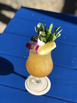 A refreshing tropical cocktail is served in a stemmed glass on a blue table. The drink features a frothy orange concoction and is garnished with a sprig of fresh mint, a purple orchid flower, and lemon slices.