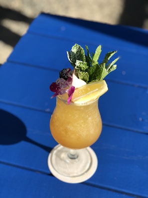 A refreshing tropical cocktail is served in a stemmed glass on a blue table. The drink features a frothy orange concoction and is garnished with a sprig of fresh mint, a purple orchid flower, and lemon slices.