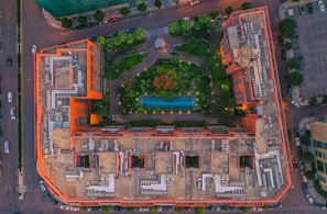 An aerial view of a large building complex with a rectangular layout, featuring a central courtyard area filled with greenery and a swimming pool. The building is bordered by streets with vehicles visible. The structure has a distinct architectural design with multiple sections and rooftop mechanical equipment.