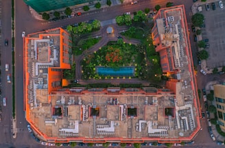 An aerial view of a large building complex with a rectangular layout, featuring a central courtyard area filled with greenery and a swimming pool. The building is bordered by streets with vehicles visible. The structure has a distinct architectural design with multiple sections and rooftop mechanical equipment.