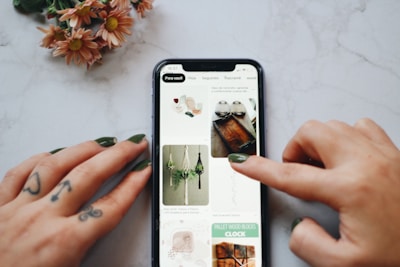 A person's hands, with tattoos and painted nails, are interacting with a smartphone displaying a Pinterest-like interface on a marble surface. The screen shows images of crafts and DIY projects including macrame plant hangers and wooden trays. A small bouquet of flowers with pink petals lies nearby.