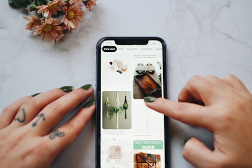 A person's hands, with tattoos and painted nails, are interacting with a smartphone displaying a Pinterest-like interface on a marble surface. The screen shows images of crafts and DIY projects including macrame plant hangers and wooden trays. A small bouquet of flowers with pink petals lies nearby.