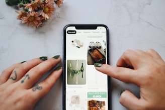 A person's hands, with tattoos and painted nails, are interacting with a smartphone displaying a Pinterest-like interface on a marble surface. The screen shows images of crafts and DIY projects including macrame plant hangers and wooden trays. A small bouquet of flowers with pink petals lies nearby.