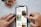 A person's hands, with tattoos and painted nails, are interacting with a smartphone displaying a Pinterest-like interface on a marble surface. The screen shows images of crafts and DIY projects including macrame plant hangers and wooden trays. A small bouquet of flowers with pink petals lies nearby.