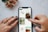 A person's hands, with tattoos and painted nails, are interacting with a smartphone displaying a Pinterest-like interface on a marble surface. The screen shows images of crafts and DIY projects including macrame plant hangers and wooden trays. A small bouquet of flowers with pink petals lies nearby.