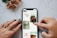 A person's hands, with tattoos and painted nails, are interacting with a smartphone displaying a Pinterest-like interface on a marble surface. The screen shows images of crafts and DIY projects including macrame plant hangers and wooden trays. A small bouquet of flowers with pink petals lies nearby.