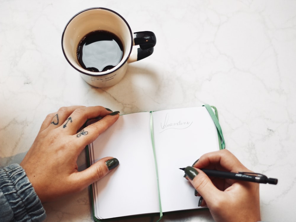 person writing on white paper beside white ceramic mug