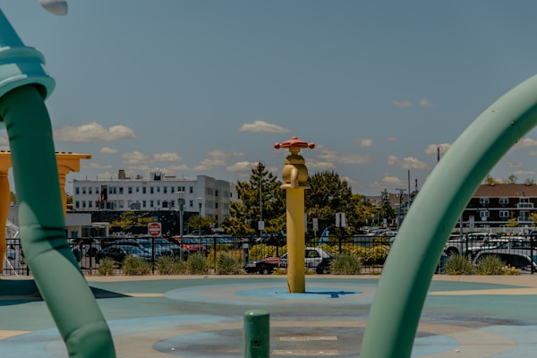 A playground water feature with colorful structures resembling a faucet and water pipe, set against an urban background with buildings and a parking lot.