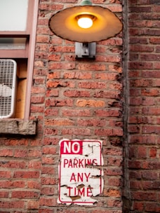 A brick wall with a weathered 'NO PARKING ANY TIME' sign hanging on it. Above the sign, there is a lit vintage-style outdoor lamp fixed to the wall. To the left of the sign, there is an air conditioning unit installed in a window.