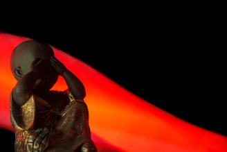 A small statue of a child monk covers its eyes. The figure is adorned in a detailed gold robe, made of a material that reflects light subtly. The background is predominantly black with a vivid red-orange wave-like element stretching diagonally, adding a dramatic contrast.