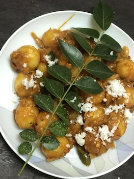 A plate of food featuring small round yellowish items covered in a thick reddish-brown sauce, garnished with green leaves and sprinkled white shavings.