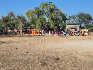 A large procession of people wearing colorful traditional clothing is moving along a dirt path. Trees and rural buildings are present in the background, and a small, brightly decorated vehicle is visible on the right side.
