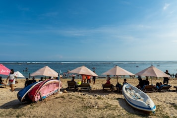 A sandy beach with multiple people relaxing under beige umbrellas. Several wooden lounge chairs are arranged facing the sea. In the foreground, a few kayaks and boats are positioned on the sand. People are swimming and engaging in water activities further out in the ocean under a clear blue sky.