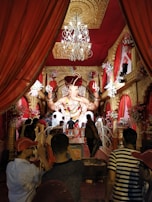 A large, intricately decorated statue of the Hindu deity Ganesha is the focal point of a beautifully adorned room, surrounded by vibrant red drapery and ornate golden details. A chandelier hangs above, and numerous people are gathered, some taking photos, creating a lively and festive atmosphere. Floral arrangements add to the colorful and celebratory setting.