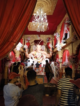A large, intricately decorated statue of the Hindu deity Ganesha is the focal point of a beautifully adorned room, surrounded by vibrant red drapery and ornate golden details. A chandelier hangs above, and numerous people are gathered, some taking photos, creating a lively and festive atmosphere. Floral arrangements add to the colorful and celebratory setting.