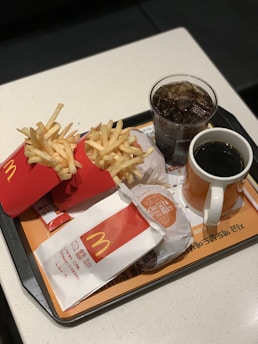 A fast food meal consisting of two packs of French fries, a wrapped burger, a glass of soda with ice, and a cup of black coffee on a tray with branded packaging.
