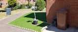 A neatly maintained front yard with an artificial grass patch bordered by stones. In the center of the grass, a potted plant with white flowers adds a decorative touch. To the right, a brown garbage bin stands next to a brick wall, accompanied by a small stone statue resembling an animal. A narrow concrete path separates the grass from a larger lawn area. The background features a row of brick houses with a garden and trees.