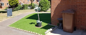 A neatly maintained front yard with an artificial grass patch bordered by stones. In the center of the grass, a potted plant with white flowers adds a decorative touch. To the right, a brown garbage bin stands next to a brick wall, accompanied by a small stone statue resembling an animal. A narrow concrete path separates the grass from a larger lawn area. The background features a row of brick houses with a garden and trees.