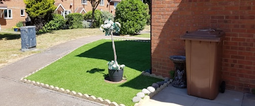A neatly maintained front yard with an artificial grass patch bordered by stones. In the center of the grass, a potted plant with white flowers adds a decorative touch. To the right, a brown garbage bin stands next to a brick wall, accompanied by a small stone statue resembling an animal. A narrow concrete path separates the grass from a larger lawn area. The background features a row of brick houses with a garden and trees.