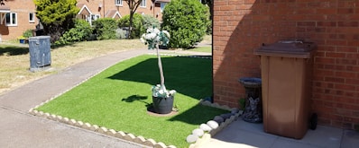 A neatly maintained front yard with an artificial grass patch bordered by stones. In the center of the grass, a potted plant with white flowers adds a decorative touch. To the right, a brown garbage bin stands next to a brick wall, accompanied by a small stone statue resembling an animal. A narrow concrete path separates the grass from a larger lawn area. The background features a row of brick houses with a garden and trees.