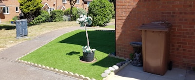 A neatly maintained front yard with an artificial grass patch bordered by stones. In the center of the grass, a potted plant with white flowers adds a decorative touch. To the right, a brown garbage bin stands next to a brick wall, accompanied by a small stone statue resembling an animal. A narrow concrete path separates the grass from a larger lawn area. The background features a row of brick houses with a garden and trees.