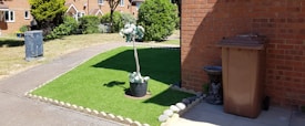 A neatly maintained front yard with an artificial grass patch bordered by stones. In the center of the grass, a potted plant with white flowers adds a decorative touch. To the right, a brown garbage bin stands next to a brick wall, accompanied by a small stone statue resembling an animal. A narrow concrete path separates the grass from a larger lawn area. The background features a row of brick houses with a garden and trees.