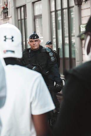 A uniformed police officer stands alert in a city environment, surrounded by civilians. The focus is on the officer, who wears a black tactical uniform and hat, holding a baton. People are in the foreground, slightly out of focus, with a city street setting in the background.