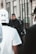 A uniformed police officer stands alert in a city environment, surrounded by civilians. The focus is on the officer, who wears a black tactical uniform and hat, holding a baton. People are in the foreground, slightly out of focus, with a city street setting in the background.