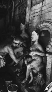 A person is sculpting a figure, applying details to a partially finished statue. The setting appears to be an artisan's workshop with various sculptures visible in the background and signs of artistic activity, such as paint buckets.