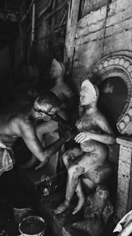 A person is sculpting a figure, applying details to a partially finished statue. The setting appears to be an artisan's workshop with various sculptures visible in the background and signs of artistic activity, such as paint buckets.
