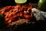 A close-up of peanuts coated in chili powder is placed on a wooden surface. Surrounding these are mounds of coarse salt and chili powder. Lime wedges are arranged in the background, adding a fresh contrast to the scene.