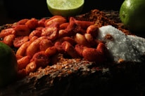 A close-up of peanuts coated in chili powder is placed on a wooden surface. Surrounding these are mounds of coarse salt and chili powder. Lime wedges are arranged in the background, adding a fresh contrast to the scene.