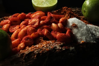 A close-up of peanuts coated in chili powder is placed on a wooden surface. Surrounding these are mounds of coarse salt and chili powder. Lime wedges are arranged in the background, adding a fresh contrast to the scene.