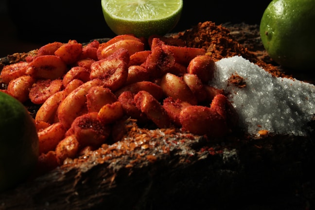 A close-up of peanuts coated in chili powder is placed on a wooden surface. Surrounding these are mounds of coarse salt and chili powder. Lime wedges are arranged in the background, adding a fresh contrast to the scene.