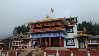 A traditional Buddhist monastery with ornate architectural details, featuring colorful decorations and flags. The building is set against a backdrop of misty trees and an overcast sky.