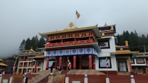 A traditional Buddhist monastery with ornate architectural details, featuring colorful decorations and flags. The building is set against a backdrop of misty trees and an overcast sky.