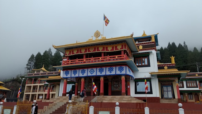 A traditional Buddhist monastery with ornate architectural details, featuring colorful decorations and flags. The building is set against a backdrop of misty trees and an overcast sky.