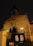 A tall, red-brick church stands against the dark night sky, illuminated by soft, warm lighting. The structure features arched windows and a cross on top of the tower. A metal fence with a gated entrance and a wooden notice board are visible in the foreground.
