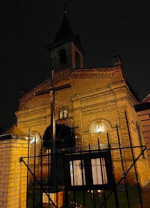 A tall, red-brick church stands against the dark night sky, illuminated by soft, warm lighting. The structure features arched windows and a cross on top of the tower. A metal fence with a gated entrance and a wooden notice board are visible in the foreground.