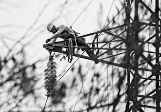 A person is climbing a metal tower, possibly an electrical or telecommunications structure, wearing safety gear and a helmet. The surrounding area appears out of focus, highlighting the lattice framework of the tower.