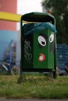 A green trash bin with cartoon eyes painted on it stands on grass, surrounded by some graffiti. A blue plastic liner is visible inside. In the background, there is a fence, a bicycle, and blurred greenery.