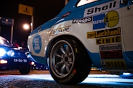 A close-up of a vintage racing car with multiple sponsor logos including Shell, Bilstein, and Dunlop. The car is illuminated under artificial lighting on a paved surface. Nearby, another vehicle with large headlights and a visible license plate is partially in view. The setting appears to be a nighttime motorsport event with signs visible in the background.