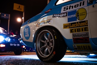 A close-up of a vintage racing car with multiple sponsor logos including Shell, Bilstein, and Dunlop. The car is illuminated under artificial lighting on a paved surface. Nearby, another vehicle with large headlights and a visible license plate is partially in view. The setting appears to be a nighttime motorsport event with signs visible in the background.