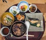 An assortment of traditional Japanese dishes arranged on a wooden tray, featuring a variety of vegetables, rice, soup, pickles, and a piece of grilled fish garnished with mustard and served on a green leaf. The presentation is colorful and includes small bowls and a cup of tea.