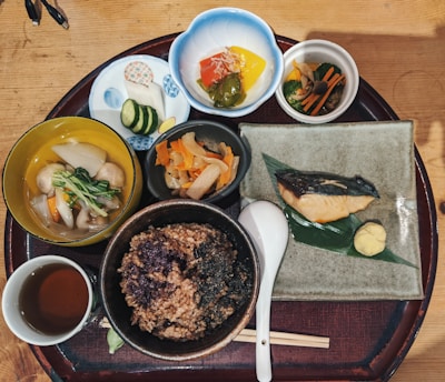 An assortment of traditional Japanese dishes arranged on a wooden tray, featuring a variety of vegetables, rice, soup, pickles, and a piece of grilled fish garnished with mustard and served on a green leaf. The presentation is colorful and includes small bowls and a cup of tea.