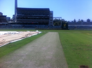 A cricket stadium with an empty pitch in the foreground, surrounded by green grass and covered areas. The stands are visible in the background, and a large structure with seating is centrally located. A scoreboard is visible in the distance.
