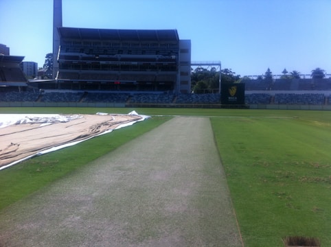A cricket stadium with an empty pitch in the foreground, surrounded by green grass and covered areas. The stands are visible in the background, and a large structure with seating is centrally located. A scoreboard is visible in the distance.