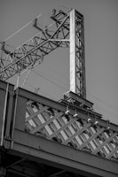 A close-up view of an industrial structure composed of metal beams and a crisscrossing lattice design, part of a larger, possibly bridge or railway infrastructure. The structure is detailed with bolts and metal ridges, complemented by power lines and an electrical component at the top.