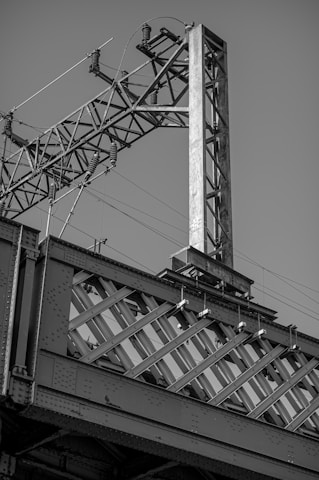 A close-up view of an industrial structure composed of metal beams and a crisscrossing lattice design, part of a larger, possibly bridge or railway infrastructure. The structure is detailed with bolts and metal ridges, complemented by power lines and an electrical component at the top.